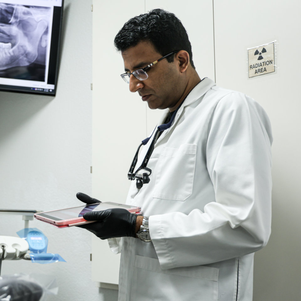 Una persona con bata de laboratorio y guantes sostiene una tableta en una sala médica con una imagen de rayos X en el monitor y un cartel de advertencia de radiación en la pared.