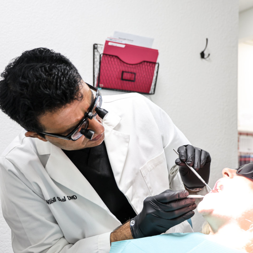 A dentist wearing magnifying glasses examines a patient's mouth in a dental clinic.