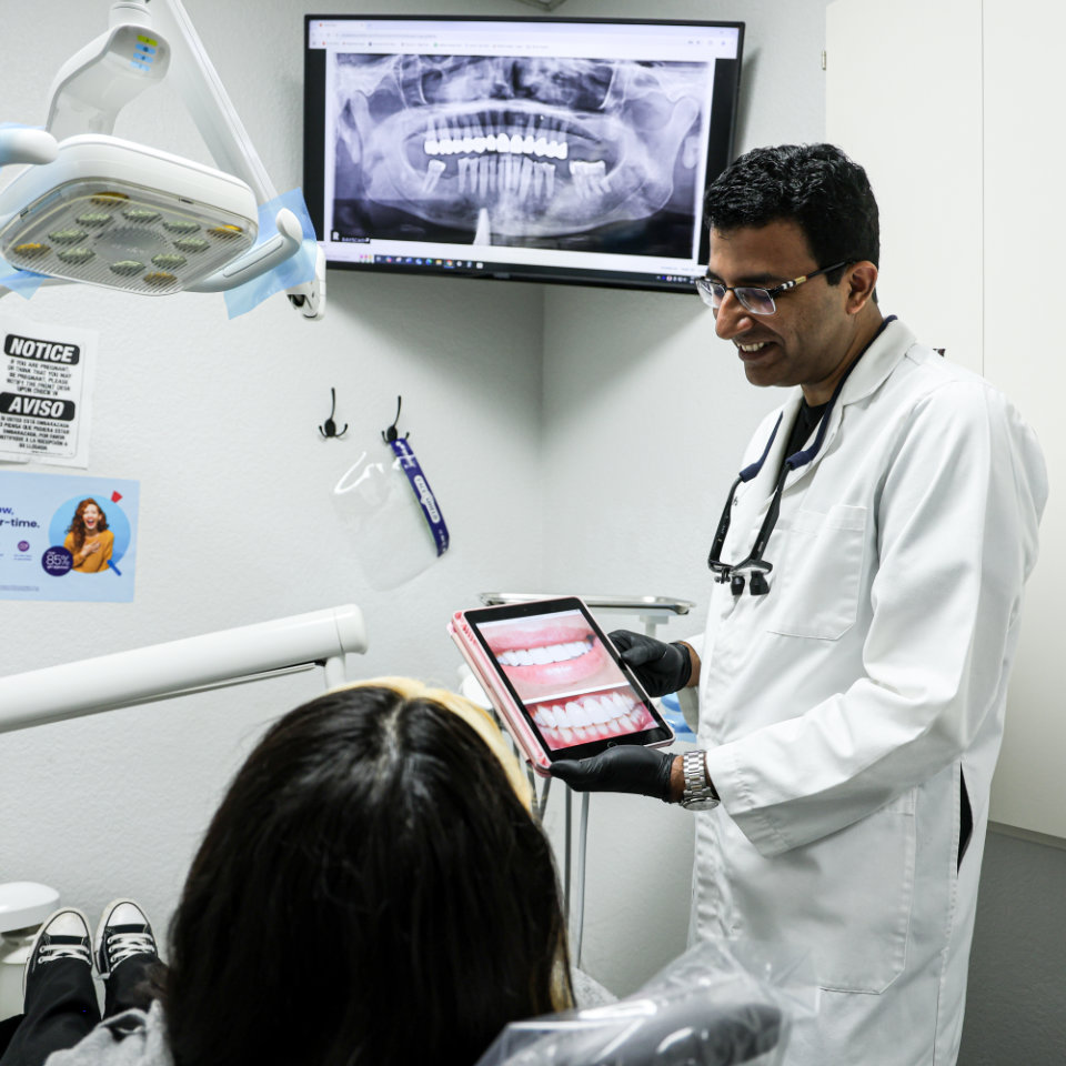 Un odontólogo muestra a un paciente dental imágenes de dientes en una tableta mientras se visualiza una radiografía dental en una pantalla en la sala de examen.