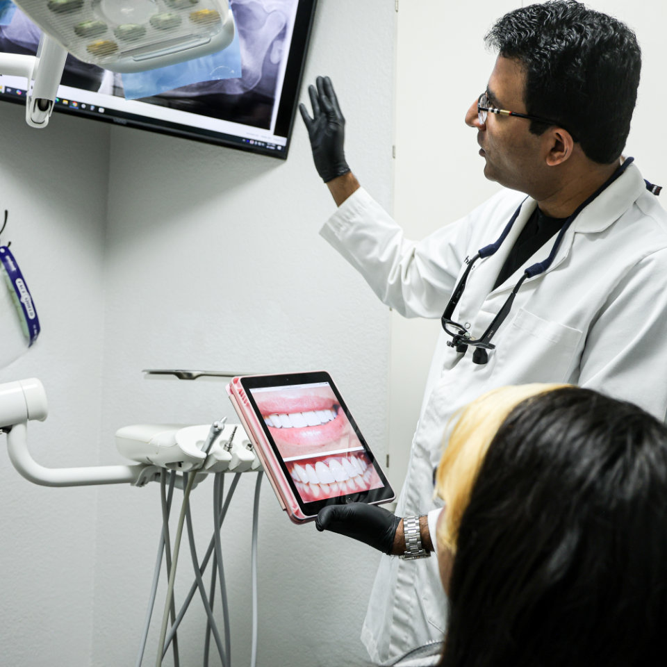 A dentist wearing gloves explains dental X-rays on a monitor to a patient while holding a tablet displaying images of teeth.