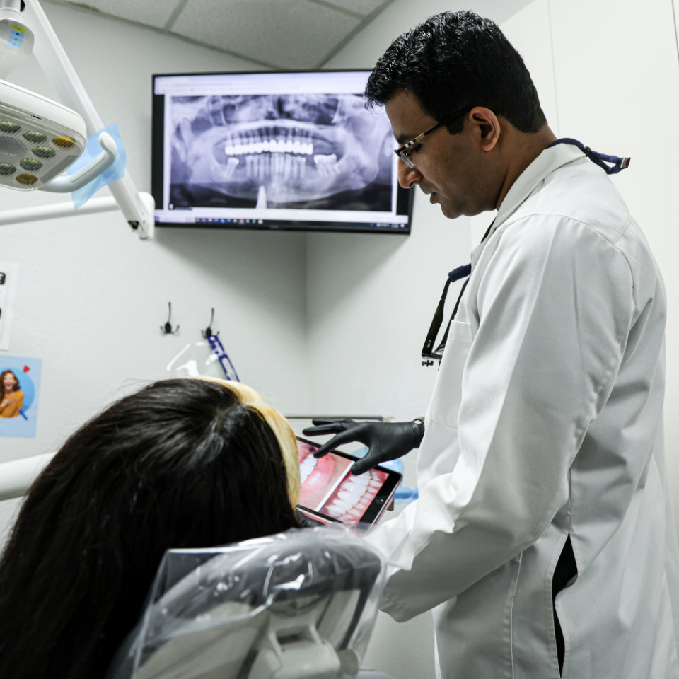 A dentist in a white coat shows a dental X-ray and mouth image to a patient seated in a dental chair in a clinic examination room.