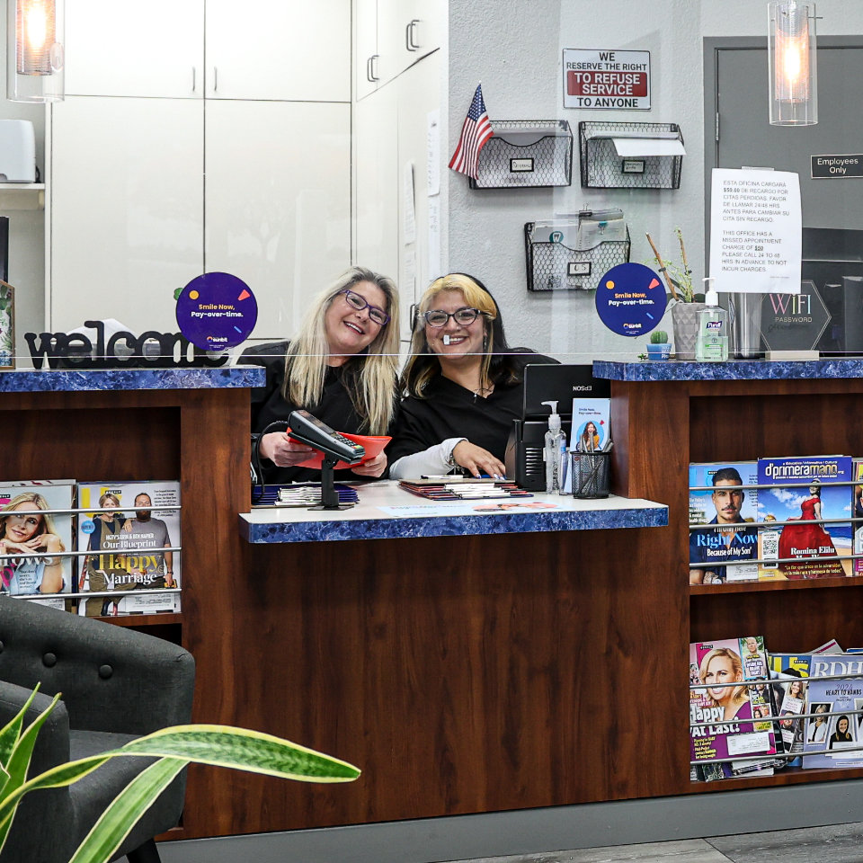 Two women sit behind a reception desk in an office, smiling at the camera. The desk has brochures, magazines, and a credit card reader. Shelves and a "No Service" sign are on the wall behind them.