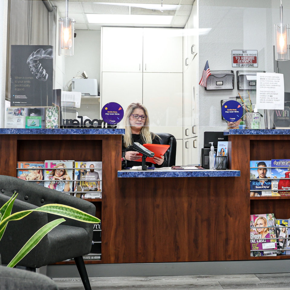 Una mujer se sienta en un escritorio de recepción leyendo una carpeta roja, rodeada de revistas, folletos y suministros de oficina en una sala de espera.