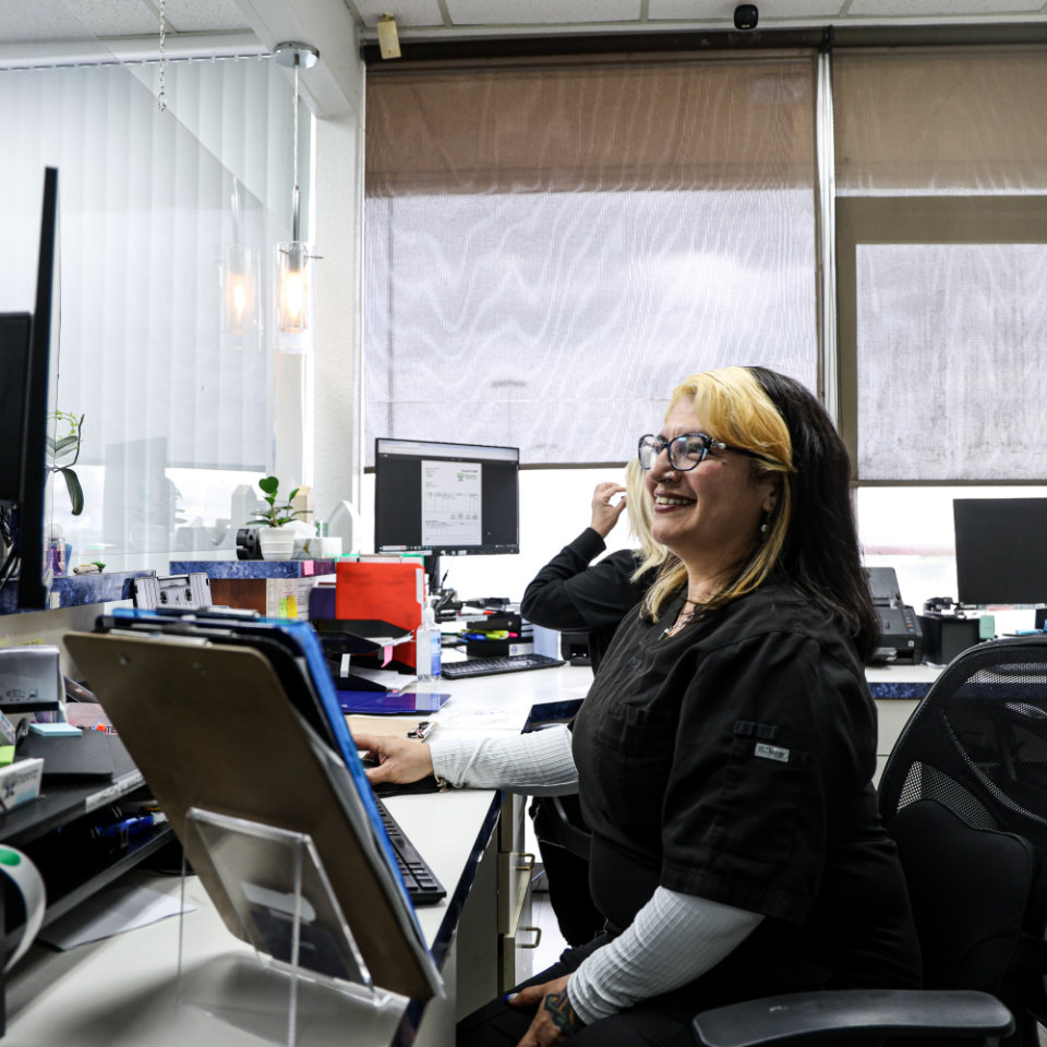 Dos empleados de oficina sentados en escritorios con computadoras. Una mujer en primer plano, con gafas y uniforme médico, sonríe mientras trabaja en su computadora.