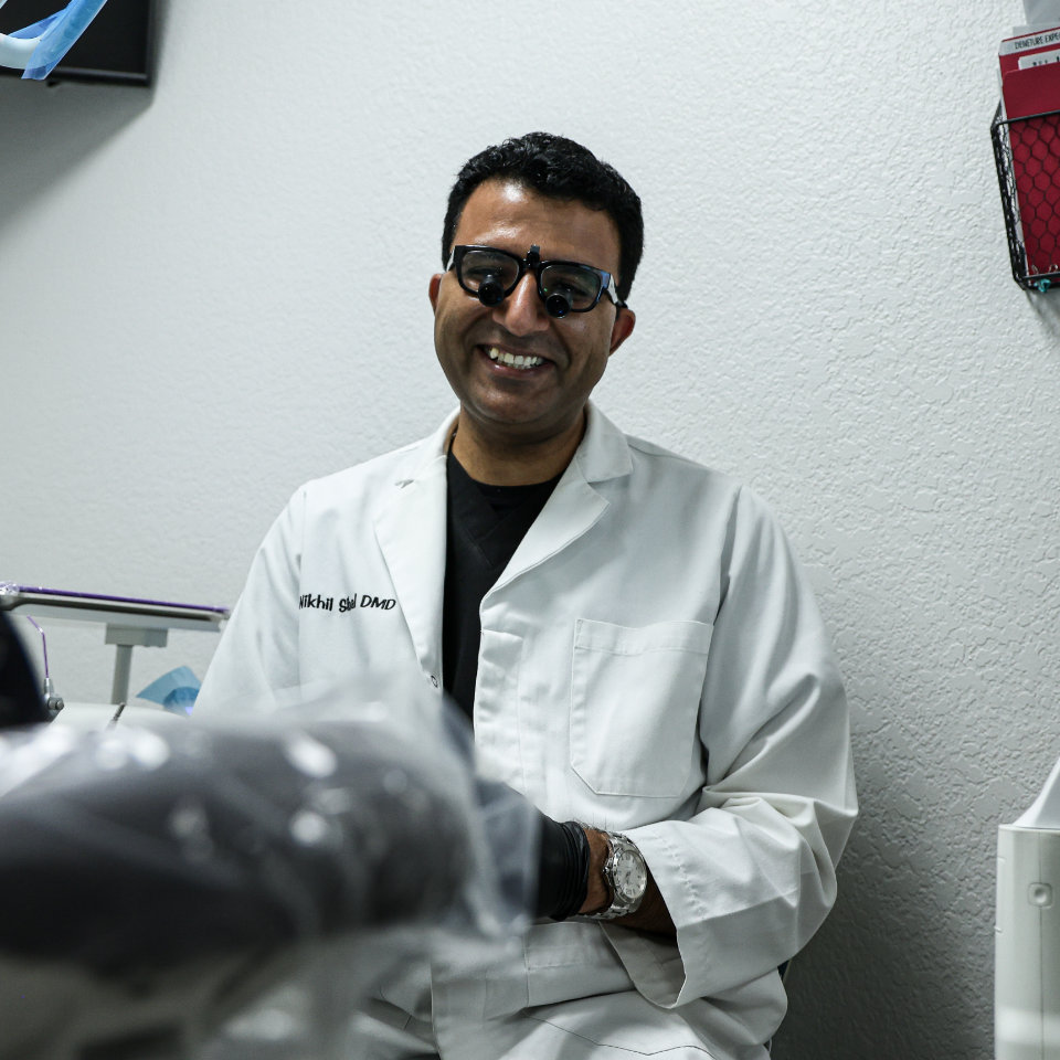 Un hombre con gafas de sol y bata blanca está sentado y sonríe en un consultorio dental.