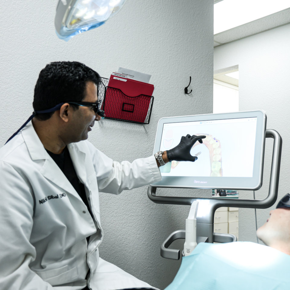 Un dentista con guantes y bata blanca señala una imagen dental en un monitor mientras examina a un paciente en una clínica.