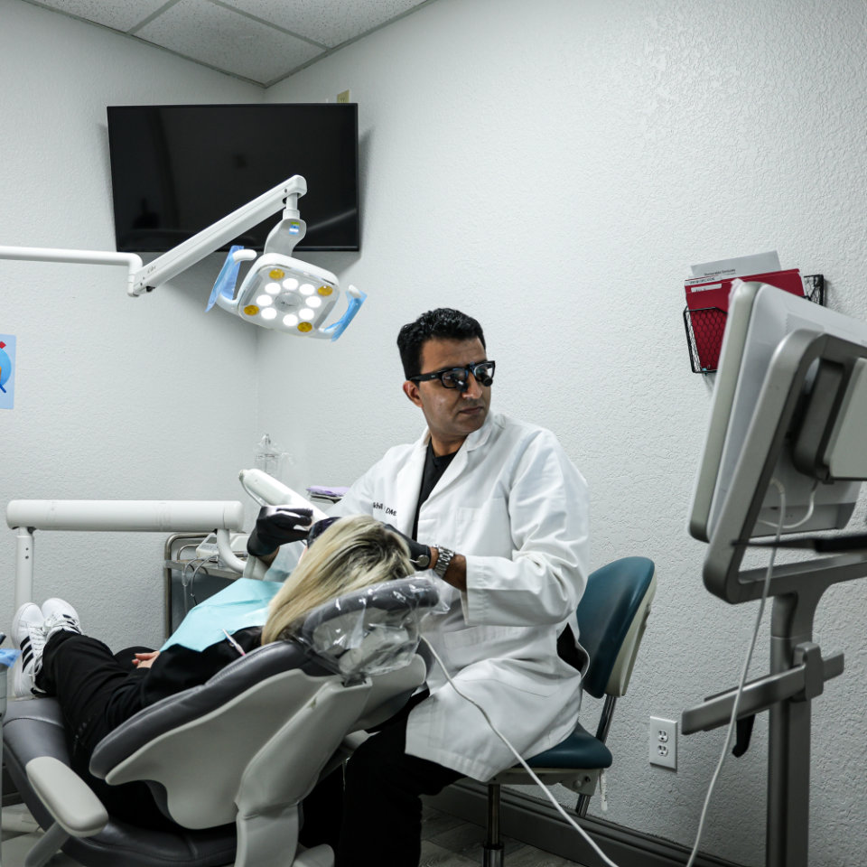 Un dentista con gafas protectoras examina a un paciente en una silla dental, con equipo dental y un monitor visibles en la sala de tratamiento.