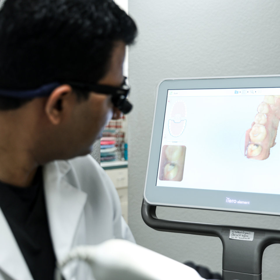 A dentist in a white coat looks at a digital dental scan displayed on a monitor showing detailed images of teeth.
