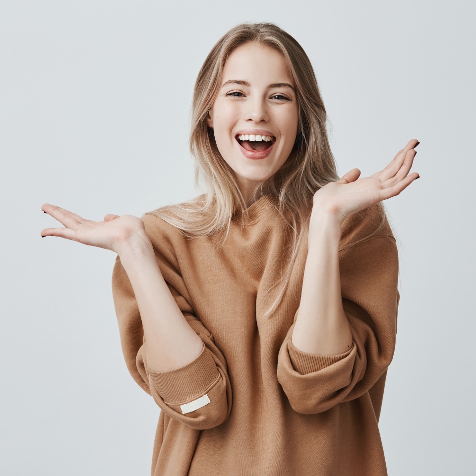 Mujer bonita con cabello largo y rubio mirando a la cámara con expresión facial emocionada y feliz, aplaudiendo con las manos contra una pared de estudio en blanco, expresando su entusiasmo con un regalo