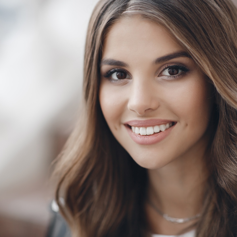 Una joven hermosa con cabello largo y ojos marrones, hermoso maquillaje, labios carnosos rosados, dulce sonrisa, morena, pasando su tiempo al aire libre en verano en la ciudad, vestida con una camiseta blanca, posando a la luz del sol. Retrato de una joven feliz. 