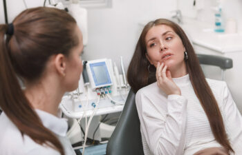 A woman sits in a dental chair touching her cheek and talking to a dentist, with dental equipment visible in the background.