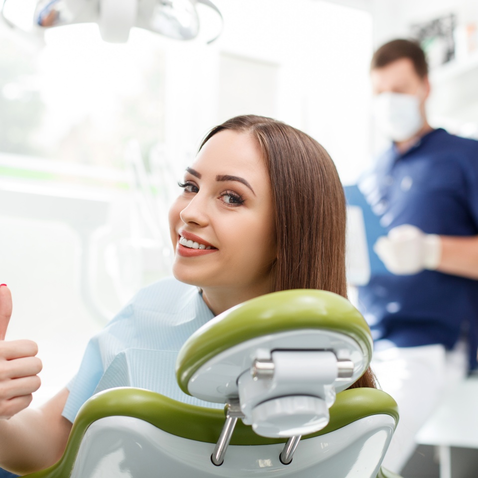 Una mujer saludable y atractiva visita a su dentista. Está sentada en la silla y mira hacia atrás con alegría. La señora levanta el pulgar y sonríe. Su dentista está de pie preparándose para el tratamiento.   