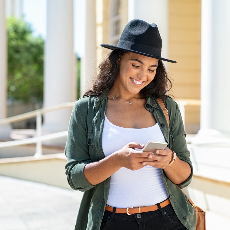Joven alegre con sombrero negro en la calle de la ciudad escribiendo un mensaje. Chica hispana a la moda navegando por internet en el teléfono al aire libre. Chica elegante feliz usando un teléfono inteligente mientras viaja por la ciudad.  