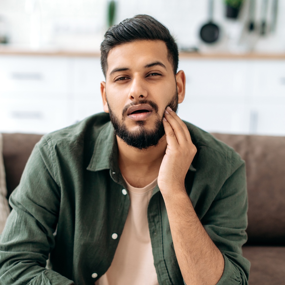 Hombre indio o árabe frustrado y poco saludable con ropa informal, presionando la mejilla con la mano, sufriendo dolor de muelas, caries, sentado en un sofá en la sala de estar, necesita la ayuda de un dentista. Concepto de problemas dentales 