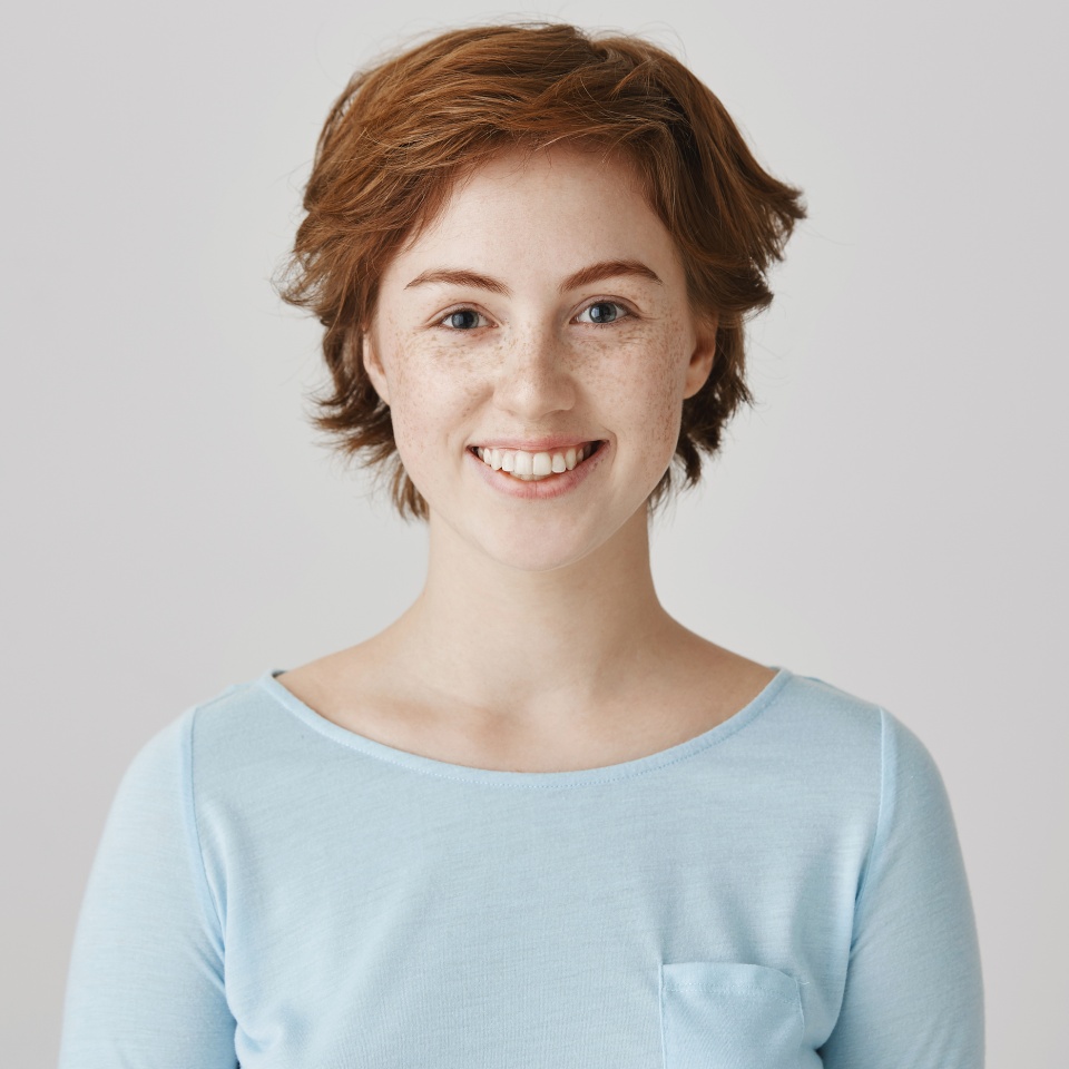 Mucho gusto. Retrato de una estudiante amigable y atractiva con cabello corto y pelirrojo, sonriendo ampliamente y mostrando una actitud positiva sobre fondo gris. Hermana que disfruta cocinar junto a su madre.  