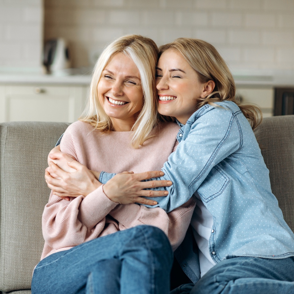 Retrato de madre caucásica y su hija. Madre madura y atractiva y su hija joven adulta, felices, sentadas en el sofá en casa, abrazándose y sonriendo. Valores familiares.  