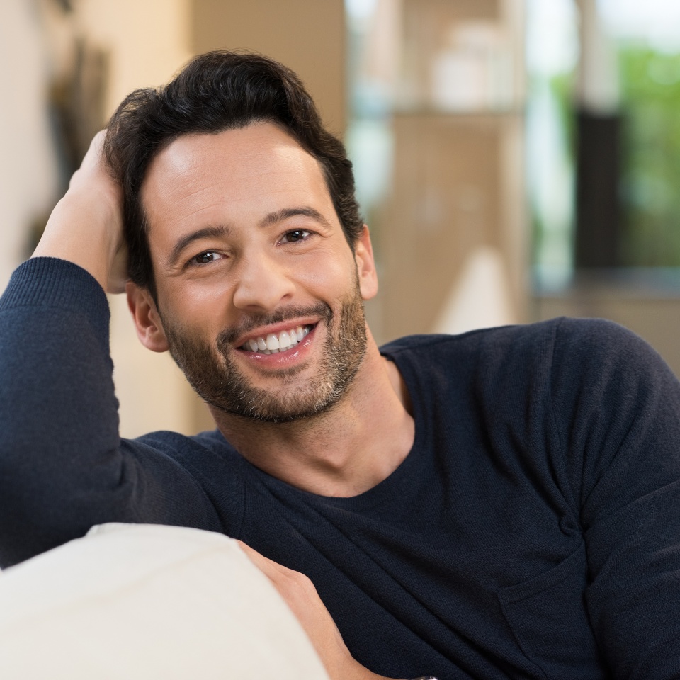 Retrato de un joven hombre sonriente y feliz descansando sentado en un sofá. Hombre atractivo mirando a la cámara. Joven latino relajándose en casa. Hombre descansando en el sofá con el brazo detrás de la cabeza.   