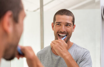 A man stands in front of a bathroom mirror brushing his teeth, wearing a grey t-shirt and smiling.