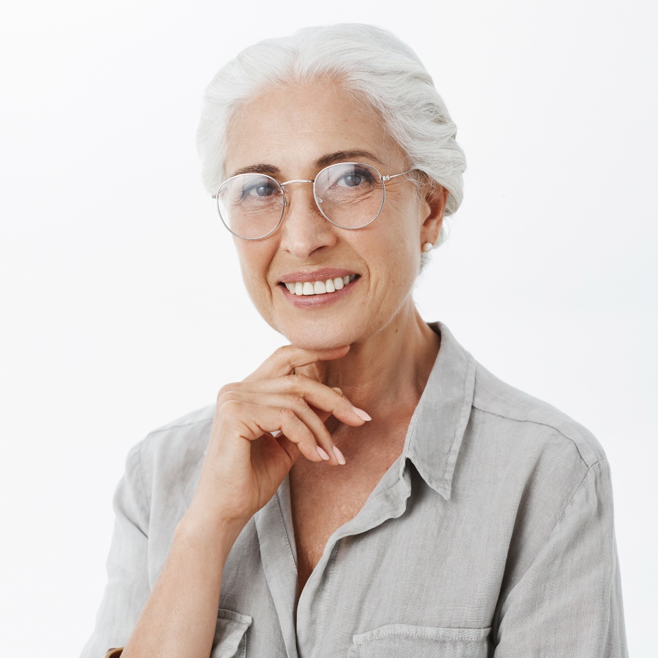 Toma de medio cuerpo de una abuela sabia y complacida, despreocupada y amable, con gafas y cabello blanco, sosteniendo la mano en la barbilla en una pose pensativa y encantada, sonriendo ampliamente con satisfacción y alegría sobre una pared gris.