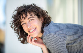 A woman with curly brown hair wearing a gray ribbed sweater smiles and rests her chin on her hand, with a blurred light background.