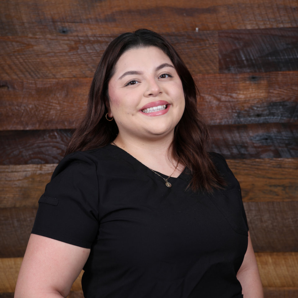 Woman with medium-length brown hair wearing a black shirt, smiling and standing in front of a wooden plank wall background.