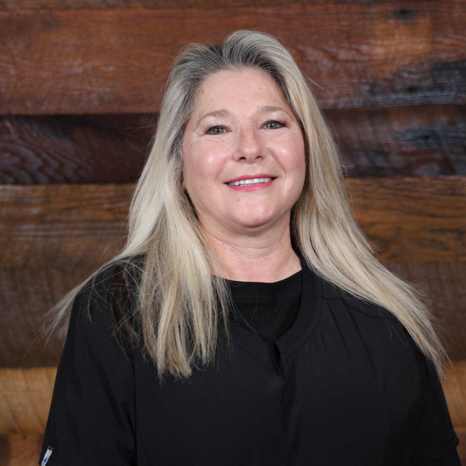 Smiling woman with long blonde hair wearing a black top, standing in front of a wooden wall background.