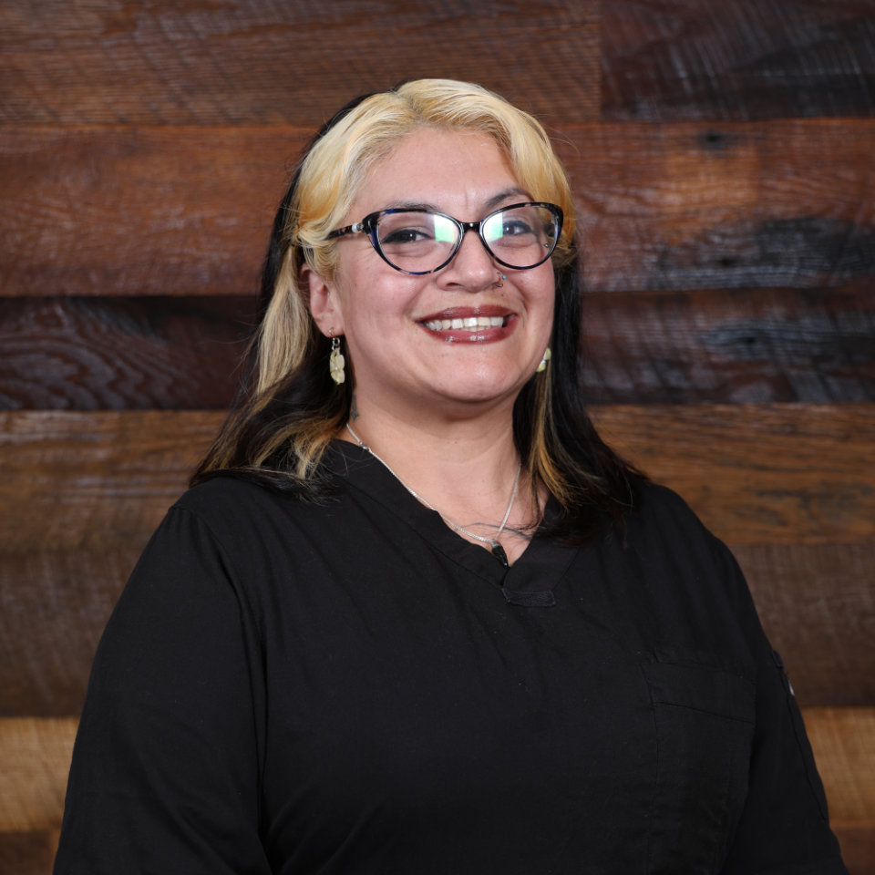 A person with blonde and black hair, glasses, and a black top smiles while standing in front of a wooden background.