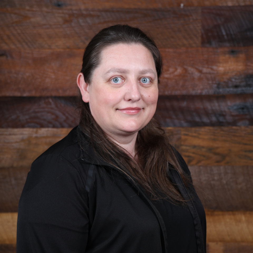 A woman with long brown hair and blue eyes, wearing a black top, stands in front of a wooden plank wall, looking at the camera with a neutral expression.