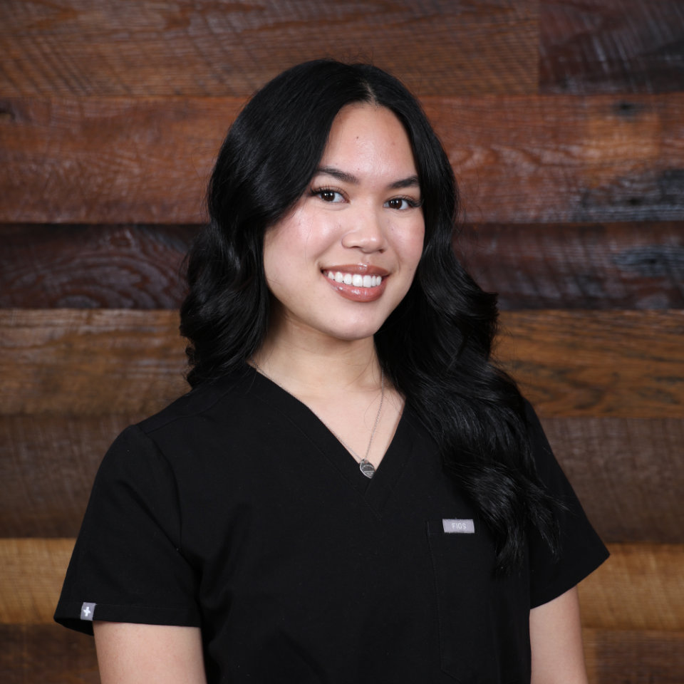 A woman with long black hair, wearing a black scrub top, stands in front of a wooden wall and smiles at the camera.