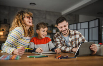 Three people sit at a table smiling while using a tablet and drawing with colored pencils and paper in a well-lit room.