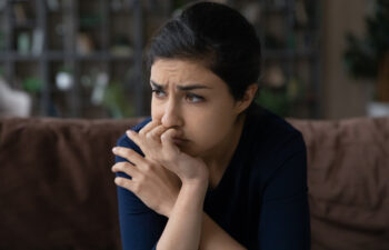 A woman sits on a couch with a worried expression, resting her chin on her hand and looking to the side.