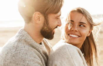 A smiling couple wearing cozy sweaters sits close together outdoors, looking at each other with joy. Sunlight shines warmly around them, creating a soft and happy atmosphere.