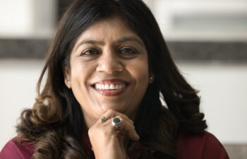 Close facial portrait of happy mature Indian woman with wavy black hair looking at camera, smiling, showing healthy white teeth, touching face, chin, promoting dental care. Cropped head shot, closeup